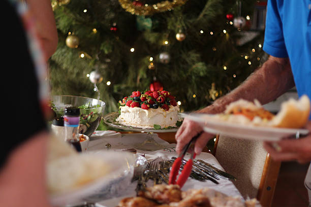 This photo shows a casual Australian Christmas lunch. A Barbecue lunch featuring charcoal grilled chicken, leg of ham, salads and dessert. In the foreground of the photo an elderly man helping himself to the buffet. The mighty pavlova is in focus with its cream, and berry topping. In the background you can see the Christmas tree with fairy lights and decorations.